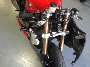 A motorcycle front end without the fairing reveals its exposed handlebar, suspension forks, and brake components—a testament to streamlined design. With a red body accented by black details, the bike rests on a light gray floor, embodying the flow and performance principles championed by Clay Green.