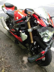 A red and black sport motorcycle, exuding flow and performance, is parked on the roadside. Its sleek design boasts a large front headlight, visible suspension, and reflective windshield. The setting includes some greenery against the paved surface.
