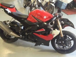 A red and black sports motorcycle with a sleek design, parked indoors. Its body features a prominent gas tank and visible metallic components, embodying the flow and performance that Clay Green coaches. In the background, another motorcycle and some equipment are partially visible.