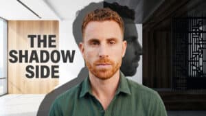 A man with short red hair and beard stands in a modern room, embodying the essence of Creating Space for Change. The words “THE SHADOW SIDE” appear beside him, while a shadowy silhouette of his face lingers behind—suggesting Daniel Ludevig’s exploration of group communication and personal transformation.