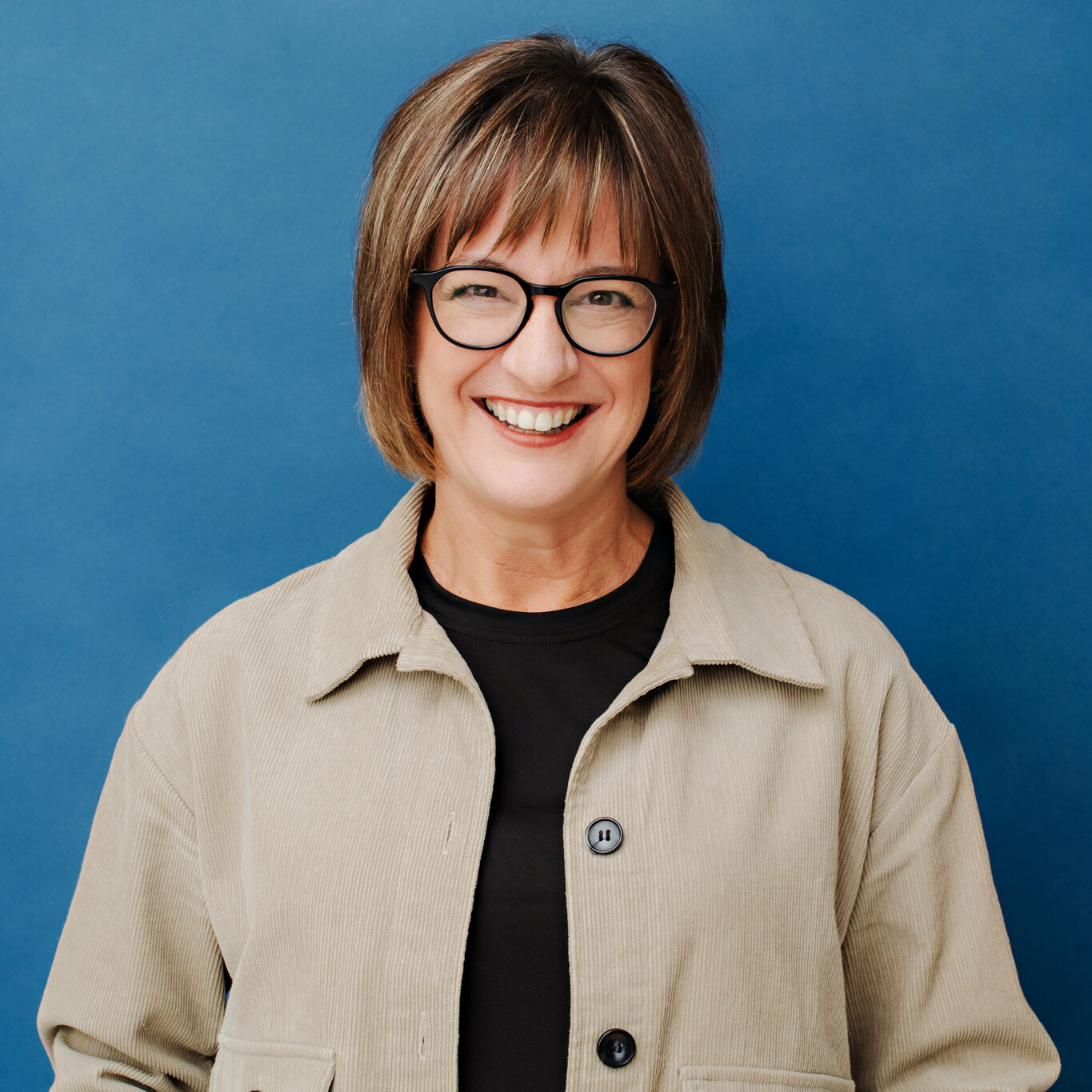 A smiling person, Rebecca Sutherns, wearing glasses, a beige shirt, and a black top stands confidently against a blue background, embodying the spirit of reinvention and career shifts that define high-achieving women.