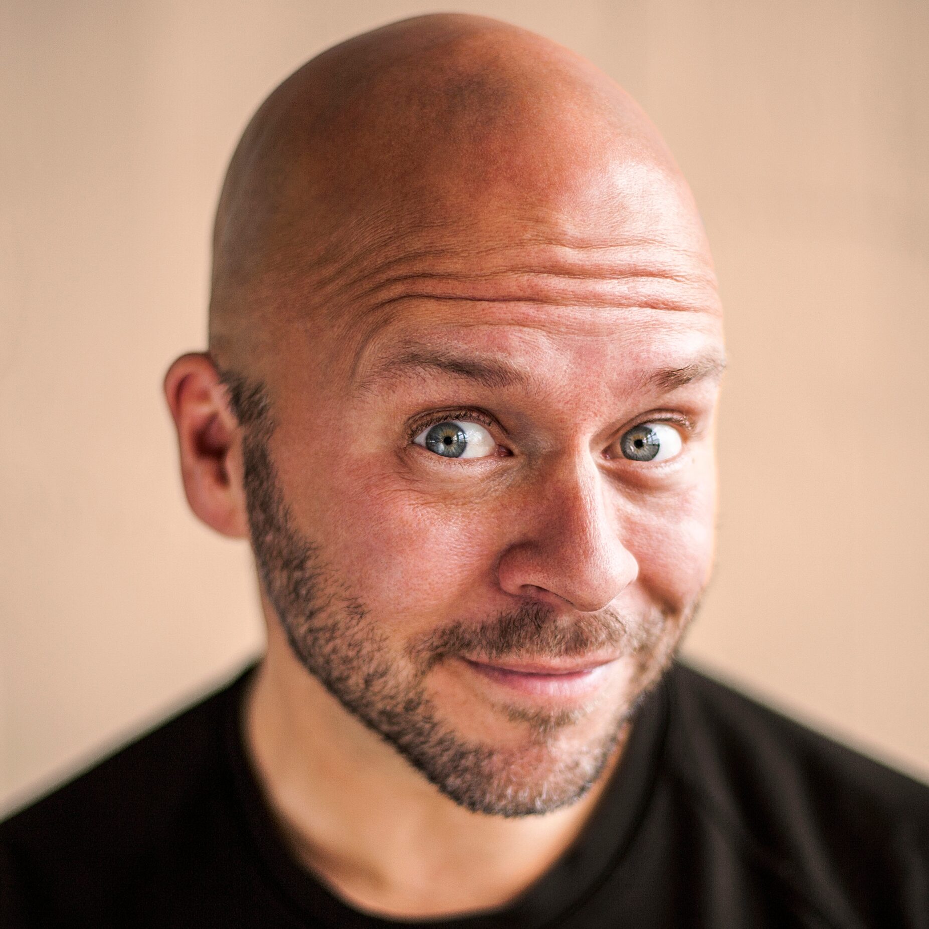 A bald man, Derek Sivers, with a trimmed beard and mustache smiles warmly, looking directly at the camera. He is wearing a black shirt and is posed against a soft, neutral background, inviting an open perspective on beliefs and emotions.