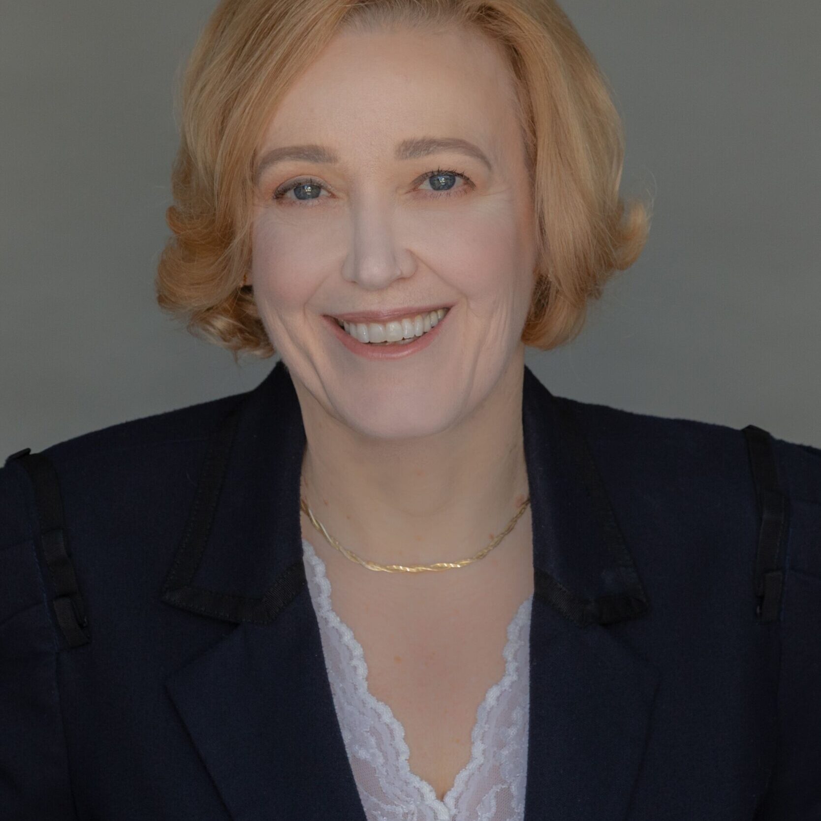 Retired Federal Judge Mary Beth O'Connor with short blonde hair, wearing a dark blazer over a white lace top and a thin gold necklace, smiles at the camera in front of a plain gray background, embodying confidence and hope in addiction recovery.