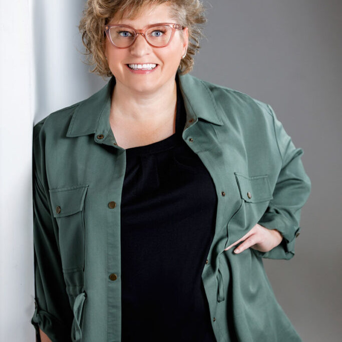 A Jennifer Jensen with curly blonde hair and glasses smiles at the camera. Channeling authentic leadership, she wears a green button-up jacket over a black top, standing against a light-colored wall with one hand on her hip.