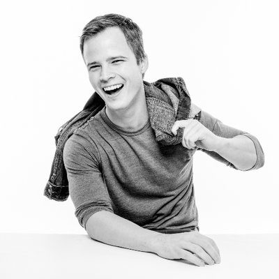 A cheerful young man from Generation Freedom, in a gray t-shirt laughing and holding a plaid shirt over his shoulder, photographed against a white background in a black and white portrait.
