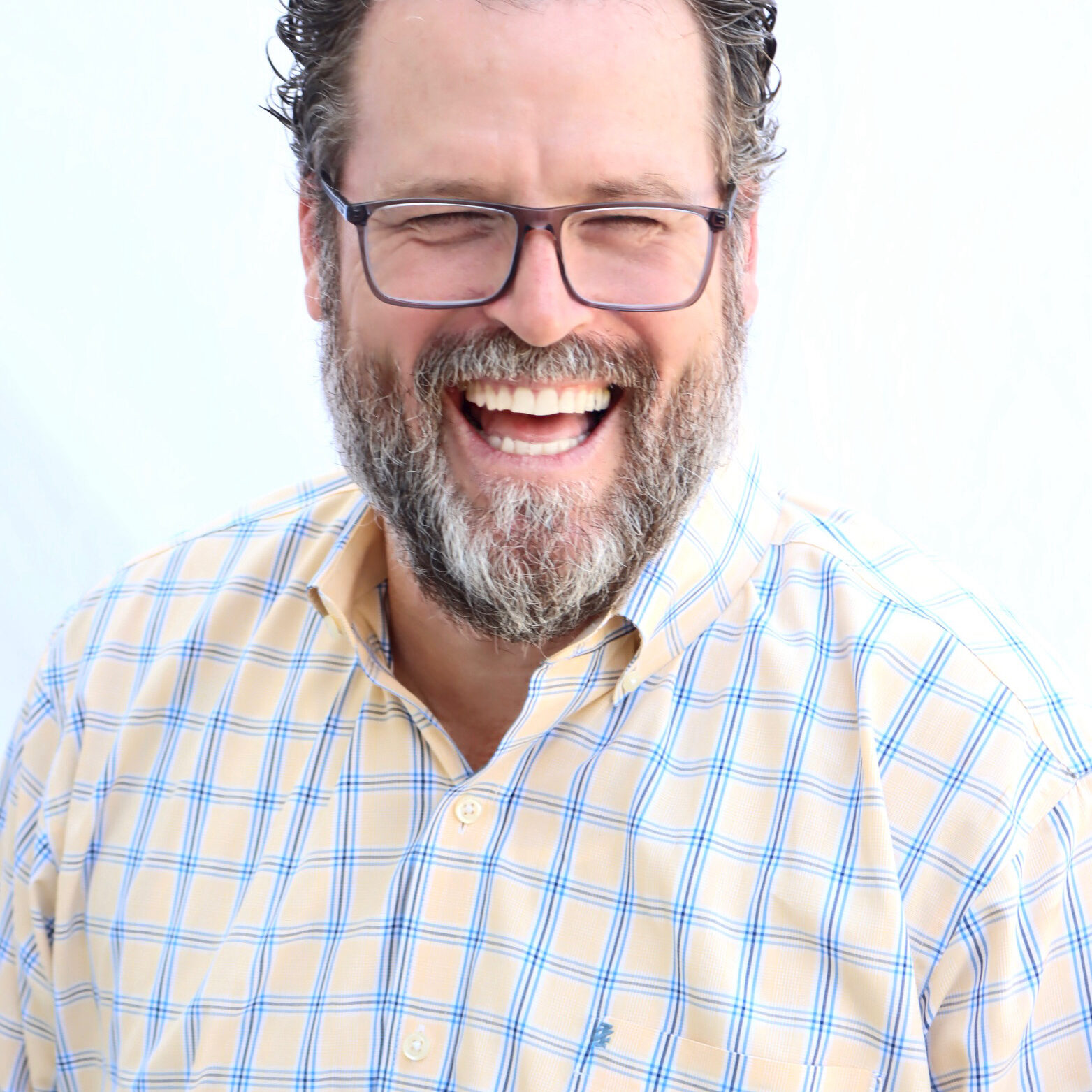 Michael Wood A bearded man with glasses, wearing a yellow and blue plaid shirt, smiles broadly and laughs while standing against a plain white background, embodying the freedom that comes from overcoming self-sabotage and outdated childhood beliefs.