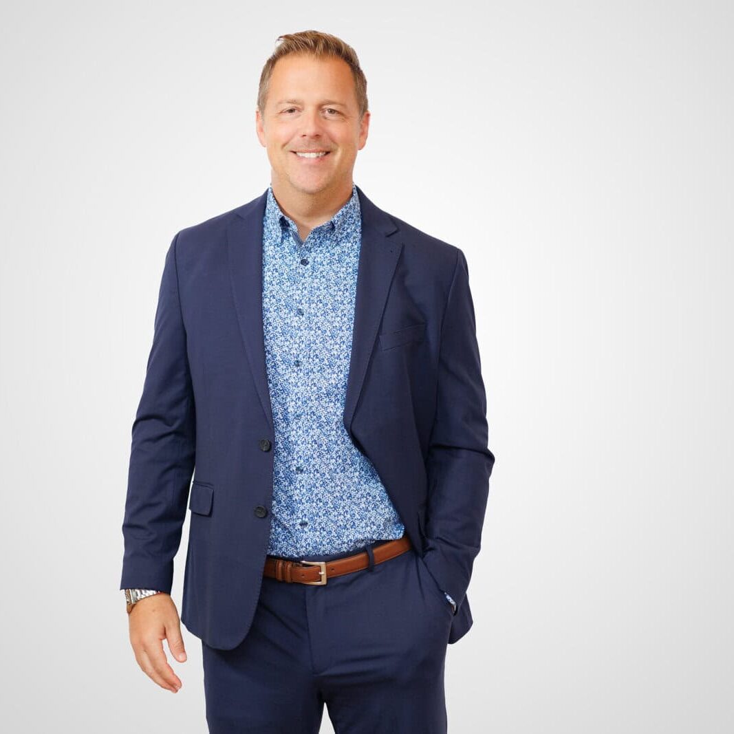 Real Estate Resilience and Long-Term Wealth: Michael Kramer's Strategic Shift. Michael Kramer, dressed in a dark blue suit and patterned shirt, stands smiling with one hand in his pocket against a plain white background.