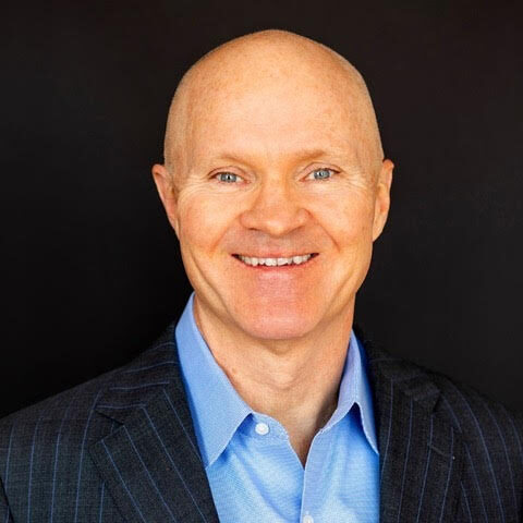 A smiling Neil Rogers, embodying a positive mindset, wears a blue dress shirt and dark pinstripe blazer as he poses in front of a plain black background.