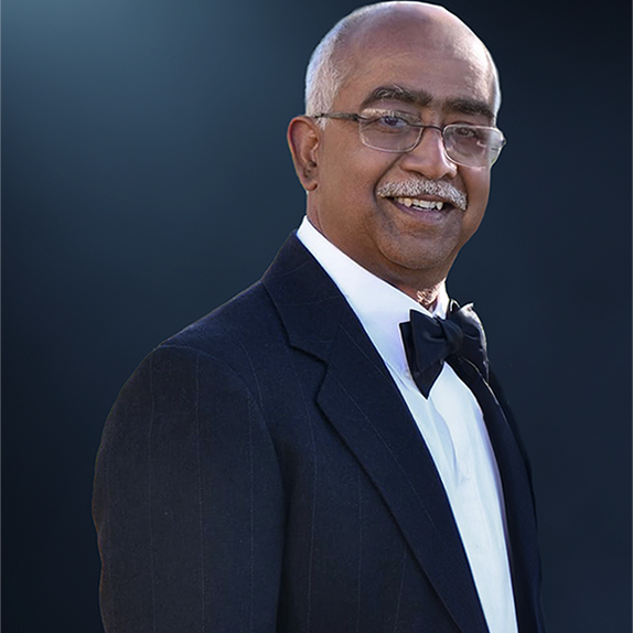 Dr. Ravi Iyer, An older man in a tuxedo with a bow tie and glasses stands against a dark background. He is smiling, with a bald head and a white mustache, exuding the distinguished charm of Dr. Ravi Iyer.