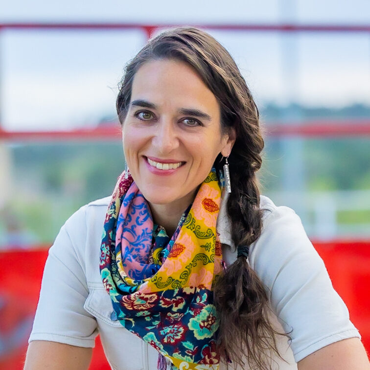 Reclaiming Life Through Adversity: Risa August’s Inspiring Tale of Personal Transformation | Ep 103. Risa August, Smiling with long brown hair and a colorful patterned scarf stands in front of a blurred red and green background. They wear a light shirt, with one braid over their shoulder, embodying self-understanding of body and taking personal responsibility for health.