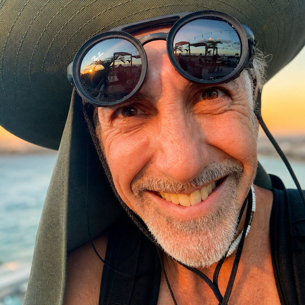 Robin Good, A smiling man with a gray beard—embracing the creator lifestyle—wears a wide-brimmed hat and round goggles reflecting a sunset. Outdoors near water, he enjoys the warm glow behind him, exuding Robin Good’s adventurous spirit.
