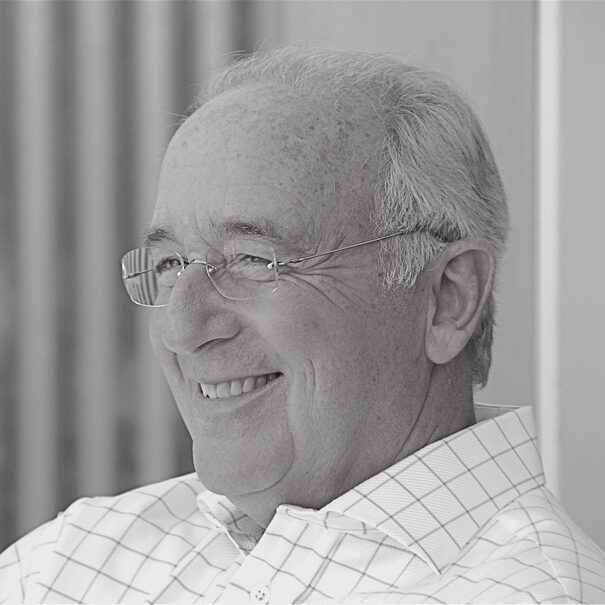 Steven Pressfield, A grayscale image of an elderly man with glasses, smiling warmly. He has short, light gray hair and is wearing a checkered shirt. The softly blurred background highlights his joyful expression, reminiscent of Steven Pressfield's themes of perseverance in "Beating Resistance.