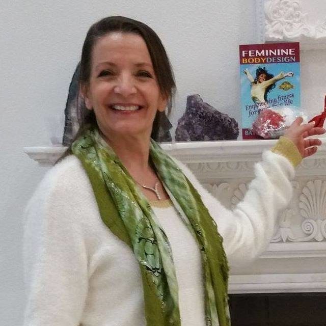 A woman with long hair and a green scarf smiles while gesturing towards Batista Gremaud's book, "Feminine Body Design," on a white mantelpiece adorned with purple crystals. The room exudes a bright and neutral decor.