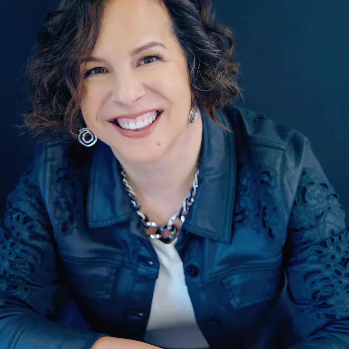 Rachel Burr, with her short curly brown hair, smiles warmly in a dark embroidered jacket, silver earrings, and a chunky necklace—a portrait capturing both elegance and transformation against a rich, dark background.