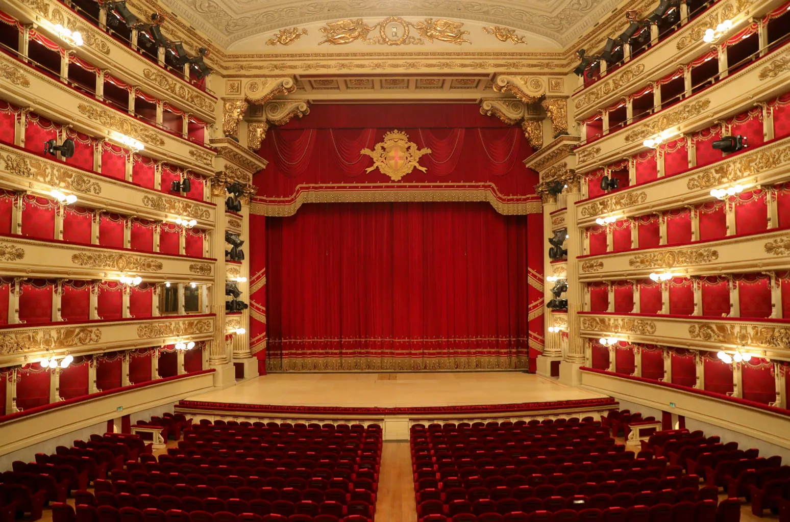 This photograph showcases the stunning interior of La Scala, the famous opera house in Milan, Italy. The grand theater is adorned in rich red and gold tones, with luxurious red velvet seats, ornate gold detailing, and tiered balconies that surround the stage. The centerpiece is the large red curtain on the stage, framed by intricate gold decorations. The overall atmosphere is one of opulence and elegance, reflecting the theater's historic significance and prestige in the world of opera.