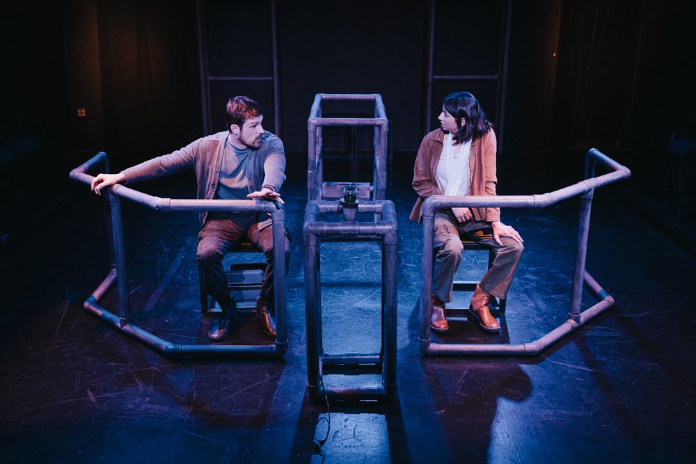 Two actors seated in a theatrical set with metal structures and dark background, engaging in a scene from 'On Our Stage Episode 6: Strange Beings'.
