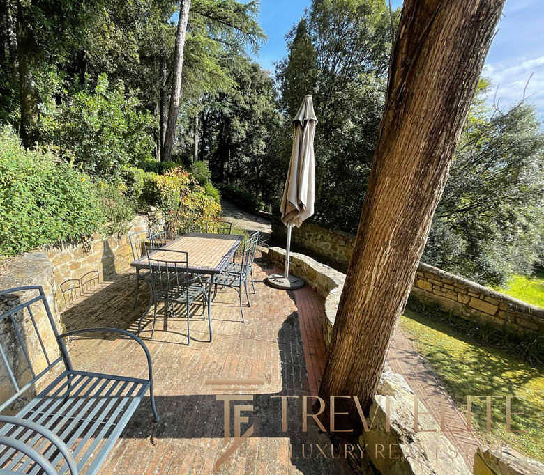 Уютная терраса на природе с деревянным столом, металлическими стульями и зонтом, окруженная зеленью и деревьями, идеально подходит для отдыха и приятных встреч на свежем воздухе.