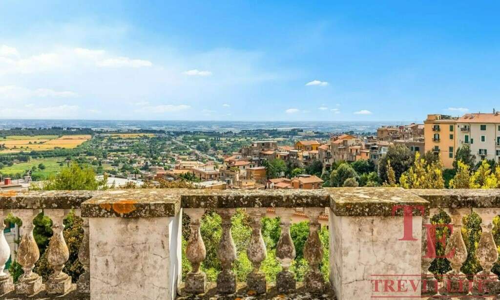 Вид на живописный итальянский пейзаж с балкона, охватывающий городские улицы, зелёные насаждения и сельскую местность на фоне голубого неба.