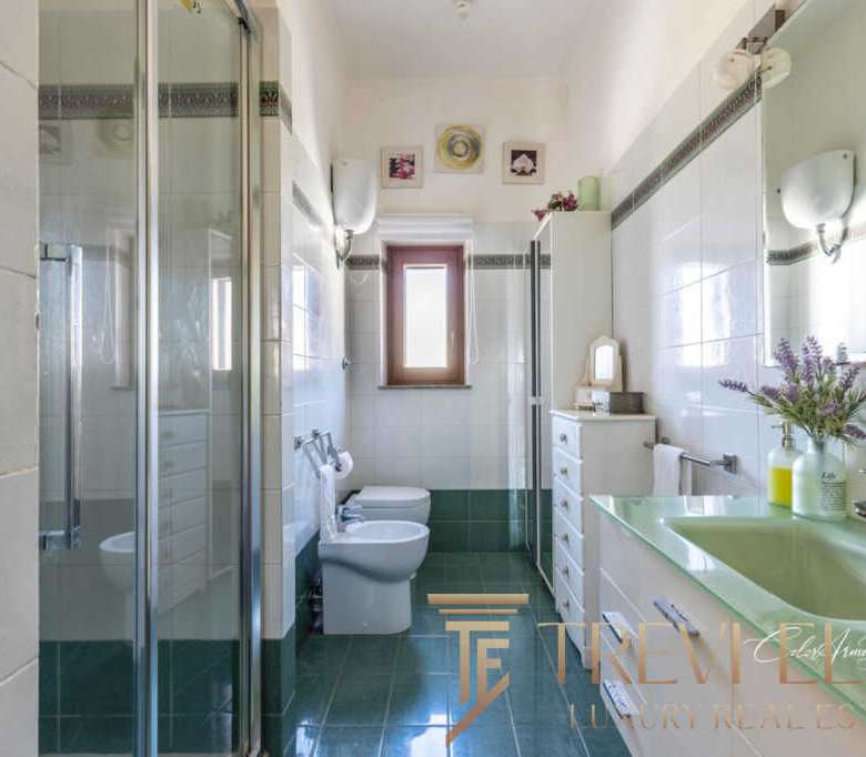 Окно ванной комнаты с зеркальным шкафом, зеленой раковиной и ванной, украшенной цветами, в светлом пространстве с современным дизайном и естественным освещением.