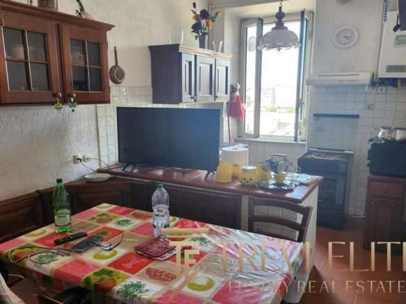 Обеденный стол в жилой кухне с яркой скатертью и напитками, рядом телевизор, открытое окно, создающее уютную атмосферу домашнего уюта и высокого уровня жизни.