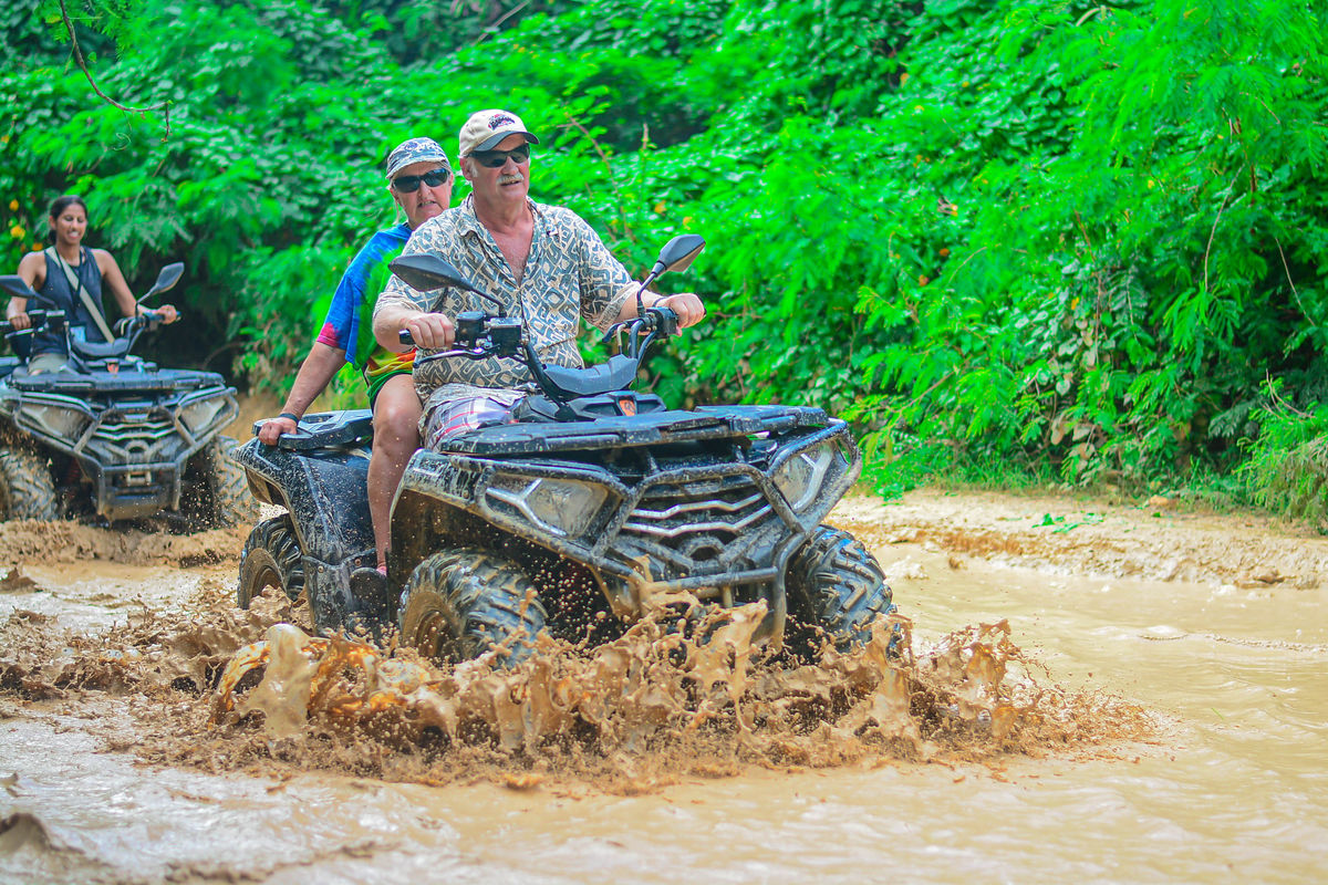 Punta Cana Two tourists riding ATVs through muddy water in lush green jungle.
