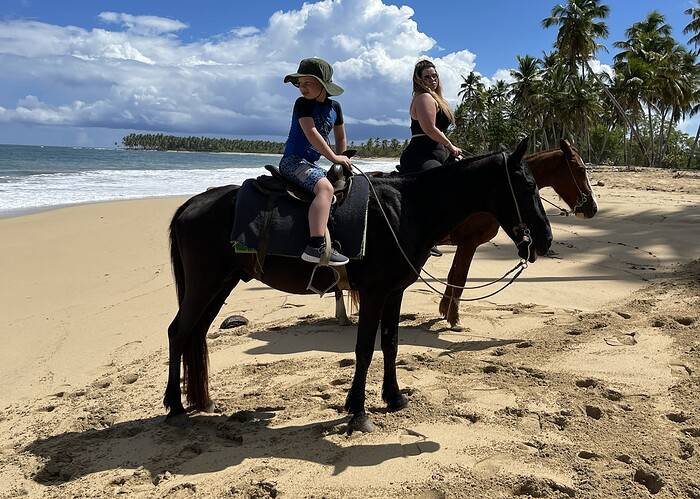 Punta Cana Two women riding horses along the sandy Punta Cana shoreline with palm trees and blue sky.