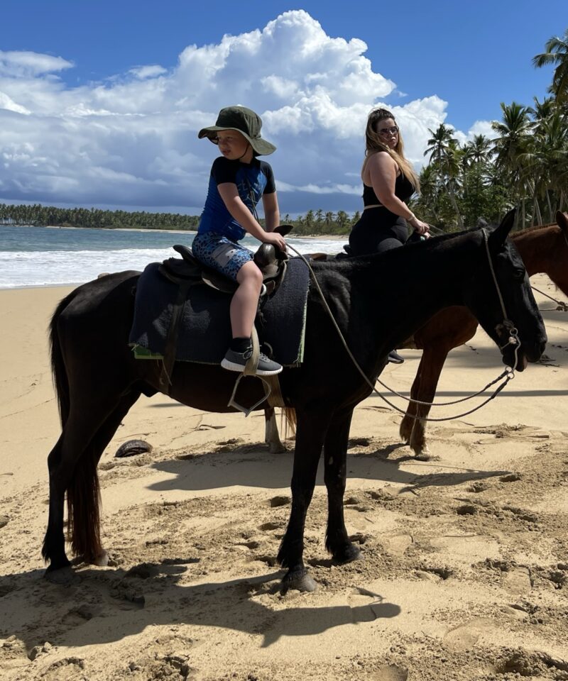 Punta Cana Two women riding horses along the sandy Punta Cana shoreline with palm trees and blue sky.