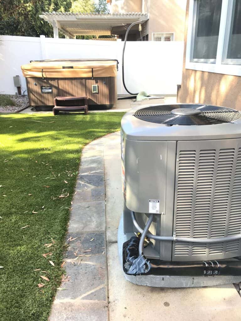 Outdoor air conditioning unit on a concrete slab next to a house with artificial grass and a hot tub in the background.