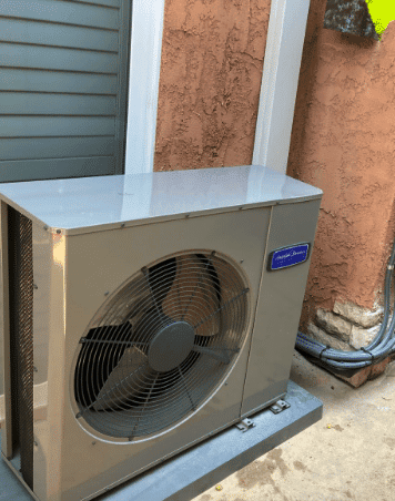 An outdoor air conditioning unit is positioned on a concrete pad next to a brown stucco wall with a vent visible above it.