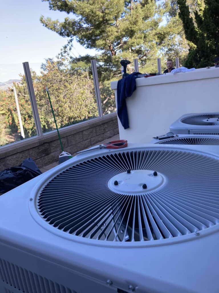 Close-up of a rooftop air conditioning unit with a fan. In the background, a person stands near a railing with trees and a clear sky visible. Tools and a broom are nearby.