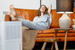 A person relaxes on a leather couch with feet up, next to a lamp on a side table, contemplating the benefits of humidifier repair while enjoying the perfectly balanced air from the nearby dehumidifier.