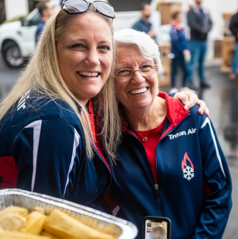 Two smiling women in matching jackets stand together at an outdoor event, embodying the spirit of "about us." The woman on the right has gray hair and glasses. In the background, another person holds a tray of food, adding to the lively atmosphere.