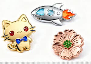 Cute gold cat enamel pin with blue bow, shiny metallic rocket ship with orange flames, and a rose gold flower pin with green accents, ideal for custom promotional products, awards, or accessories.