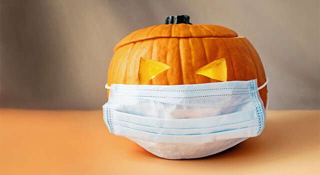 A carved pumpkin with a Halloween face wearing a blue face mask for health safety during fall festivities.