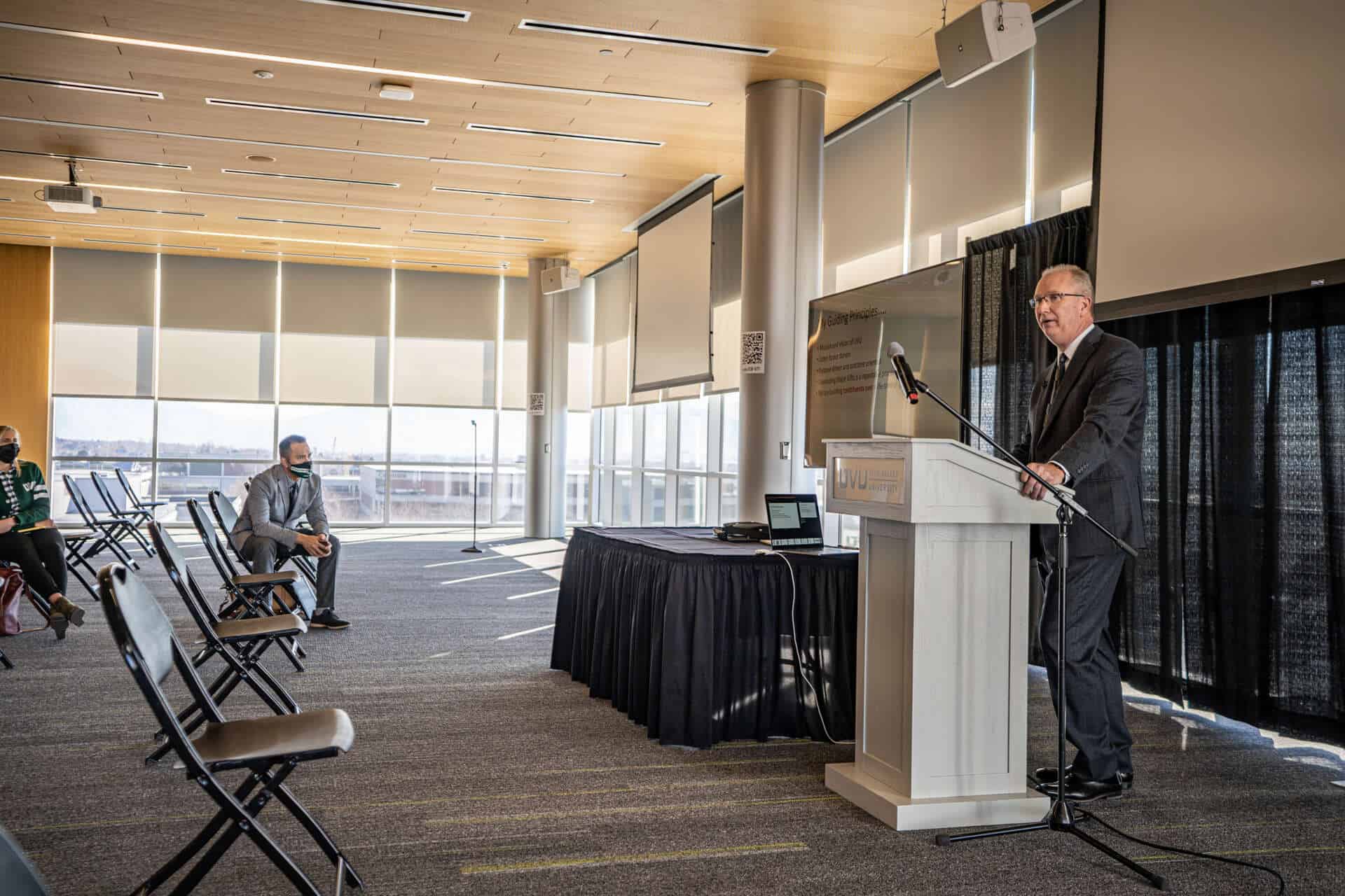 UVU Review campus presentation with speaker at podium, students seated socially distanced, modern university building interior with sunlight, for college news and student life.