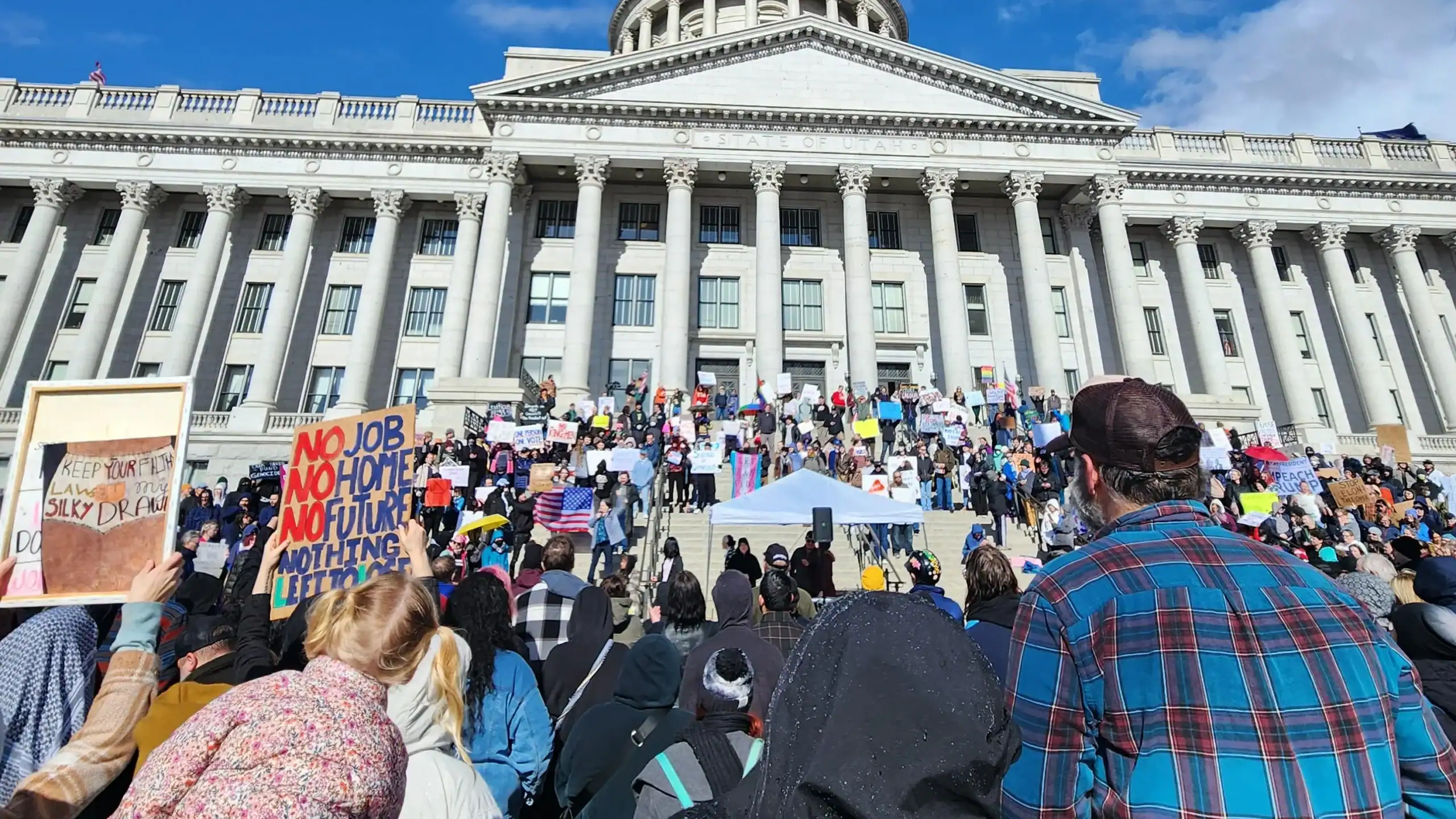 Protesters gather outside Utah State Capitol building holding signs supporting climate action and social justice. The crowd emphasizes community engagement in civic issues.