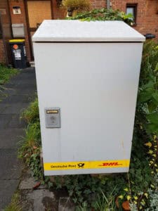 Streuhaus mit DHL- und Deutsche Post-Logo, wettergeschützt, für Paket- und Briefzustellung in Wohngebiet.
