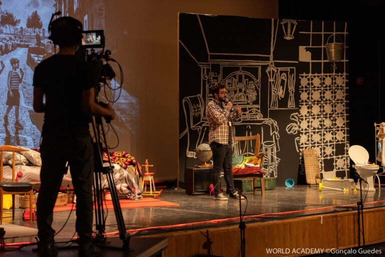 Imagem de produção teatral com apresentador no palco, equipe de gravação e cenário artístico, promovendo atividades de ensino e cultura.