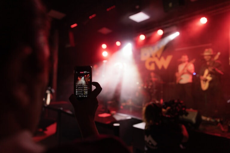 Pessoa segurando telemóvel a tirar foto em concerto no World Academy, palco de eventos culturais e musicais.