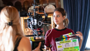 Fotografia de uma jovem atriz em gravação de filme, segurando uma claquete, durante produção cinematográfica.