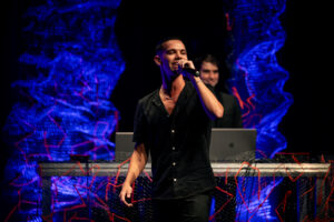 Cantor em palco com microfone e equipe de produção ao fundo, ambiente de evento musical.