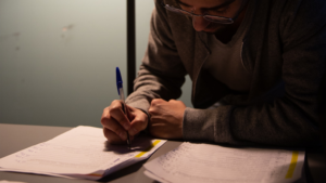 Estudante a escrever em cadernos com caneta em ambiente de estudo consagrado à formação profissional.