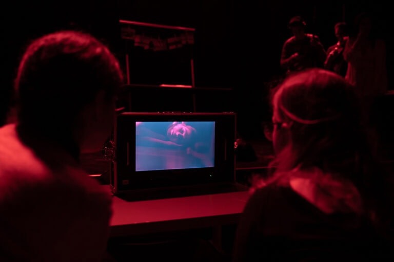Pessoas observando uma tela de monitor em ambiente escuro, com foco em tecnologias digitais educativas.