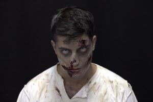 Maquiagem de terror em pessoa com aparência de zumbi, olhos brancos e expressão assustadora, fundo preto dramático.