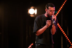 Jovem fala em palco com microfone em evento educacional.