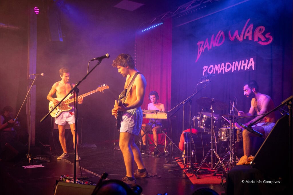 Banda Wars performando ao vivo na POMADINHA, com instrumentos musicais em palco iluminado.