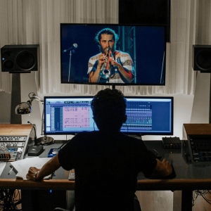 Estúdio de gravação profissional com técnico ao computador assistindo a uma performance musical na tela. Estúdio de produção musical com técnico monitorizando um vídeo de músico tocando instrumentos ao vivo.