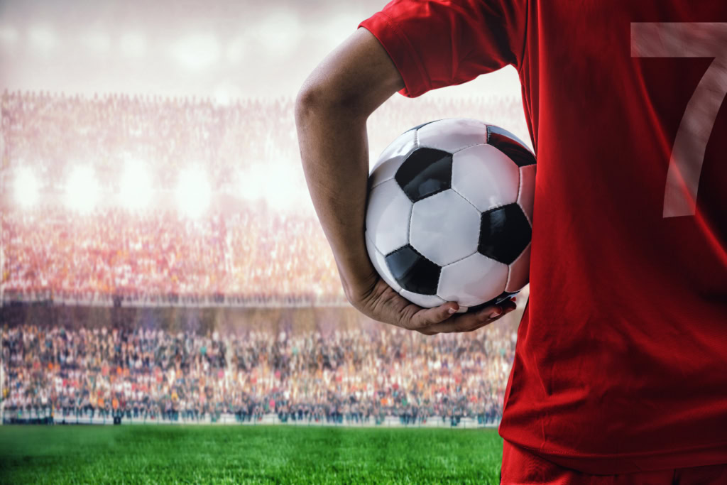 Fußballspieler mit rotem Trikot und der Nummer 7 hält einen Fußball unter dem Arm und steht auf einem Spielfeld vor einem vollen Stadion – ideal für Gamification-Software, Gewinnspiele und Wettbewerbe.