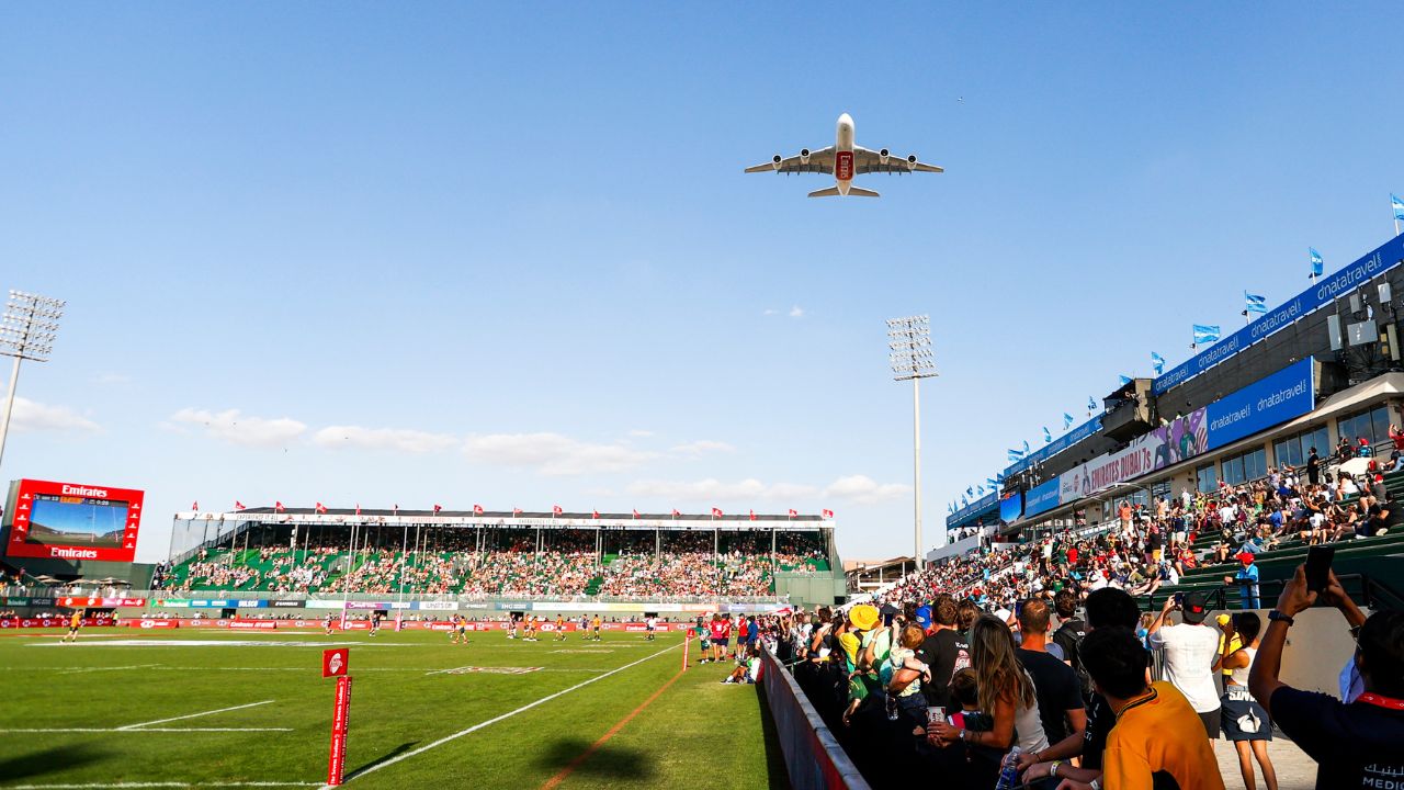 Airplane flying over Emirates Dubai 7s stadium during event.