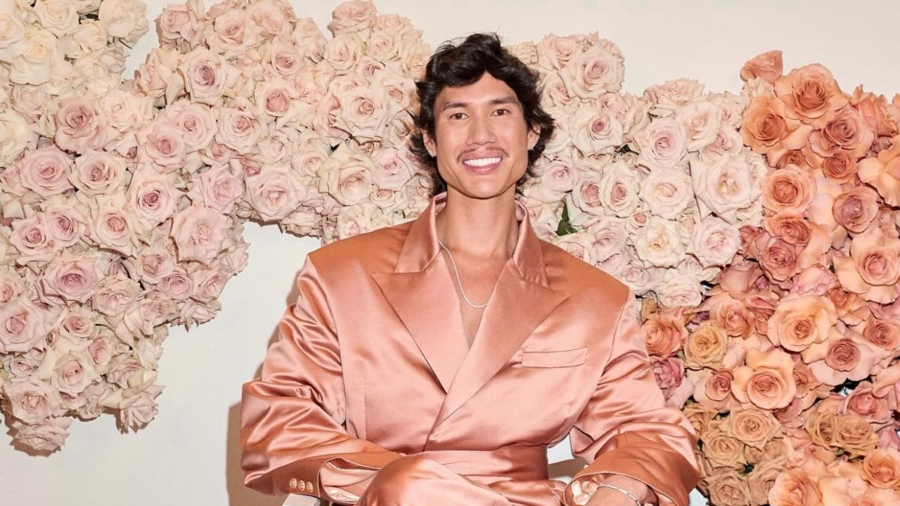 An Asian male makeup artist smiling confidently in a satin peach blazer, standing in front of a flor.